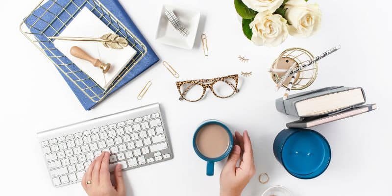 Flat lay of a stylish workspace with hands typing on a keyboard, dedicated to SEO blog writing services, including a cup of coffee, glasses, floral arrangement, books, and office supplies on a white desk