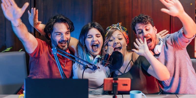 Four people are seated at a table with audio recording equipment, enthusiastically waving towards a smartphone on a tripod, suggesting they are recording or live streaming. All appear to be happy and engaged, with various types of audio gear and content repurposing tools visible.