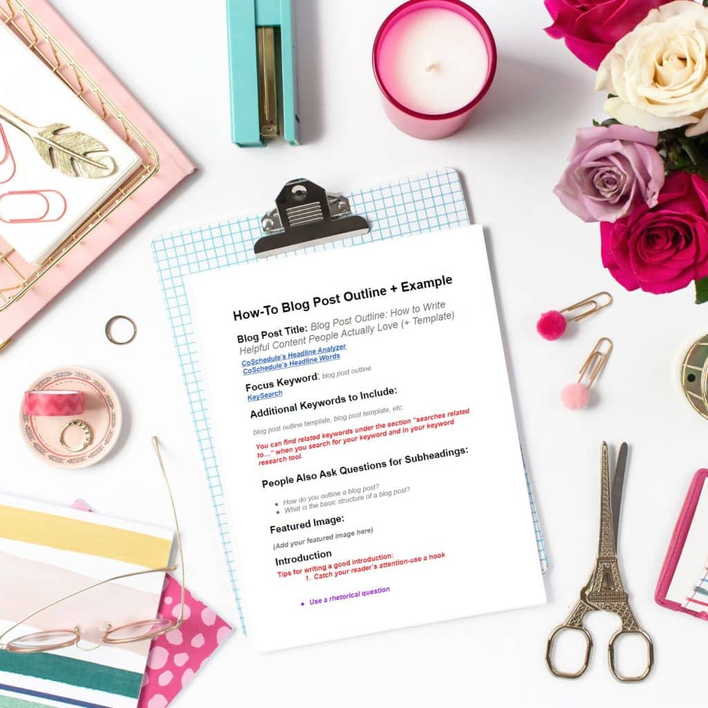 A clipboard with a printed paper titled "How-To Blog Post Outline + Example" rests on a white surface surrounded by office supplies, including a pen, pink paperclips, colorful folders, a gold scissor, a ribbon, a plant, and a bouquet of pink and white flowers.