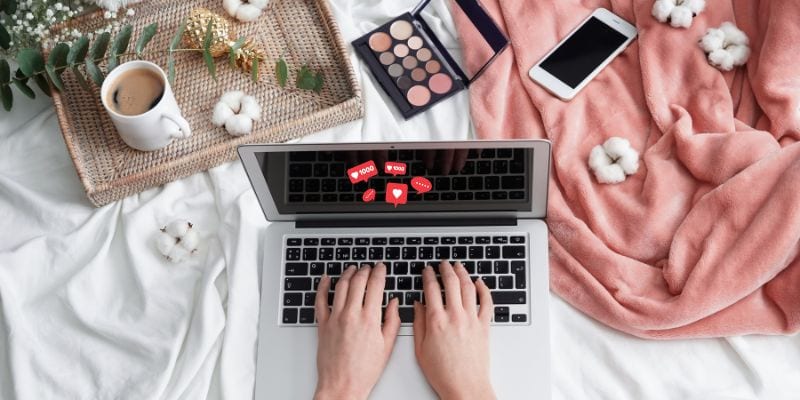 Hands typing on a laptop keyboard displaying social media icons on the screen. The laptop is surrounded by a phone, a makeup palette, a cup of coffee, cotton flowers, and a peach-colored blanket that is laying on a white surface — essentials for avoiding amateur blogging mistakes.