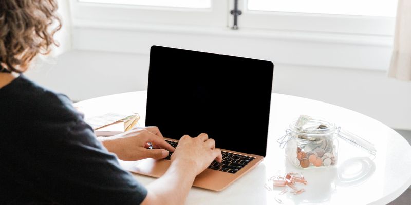 A person with curly hair is sitting at a white table typing on a laptop, possibly avoiding amateur blogging mistakes. There is a glass jar filled with coins and a notebook on the table. The background features two large windows.