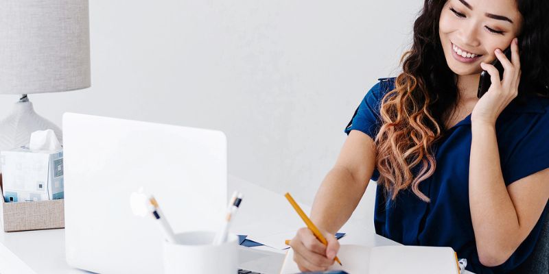 A woman in a blue shirt sits at her desk, juggling a phone call while jotting down notes with a pencil. Her laptop hums beside her, framed by a lamp and tissues. The bright room belies the blogger burnout she's battling as she strives to find balance.