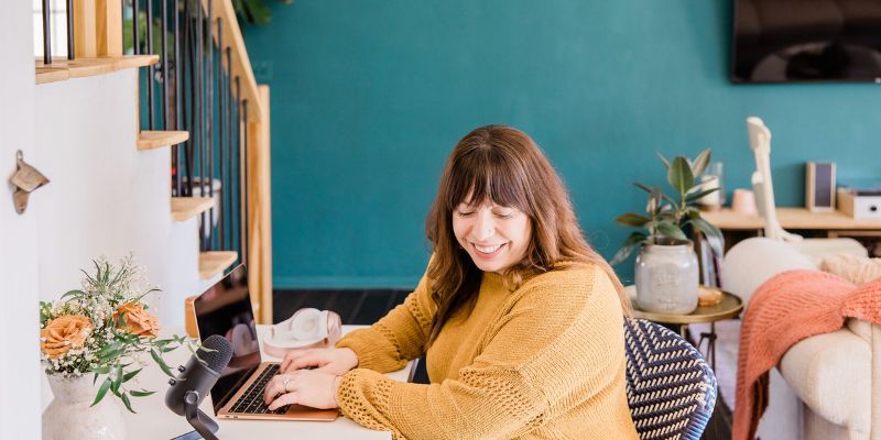 A woman with long, dark hair, wearing a yellow sweater, sits at a desk typing on a laptop. A microphone is placed on the desk, and a bouquet of flowers is on the left. She is smiling and situated in a bright room with teal walls and various decor items, balancing her passion for avoiding amateur blogging mistakes.