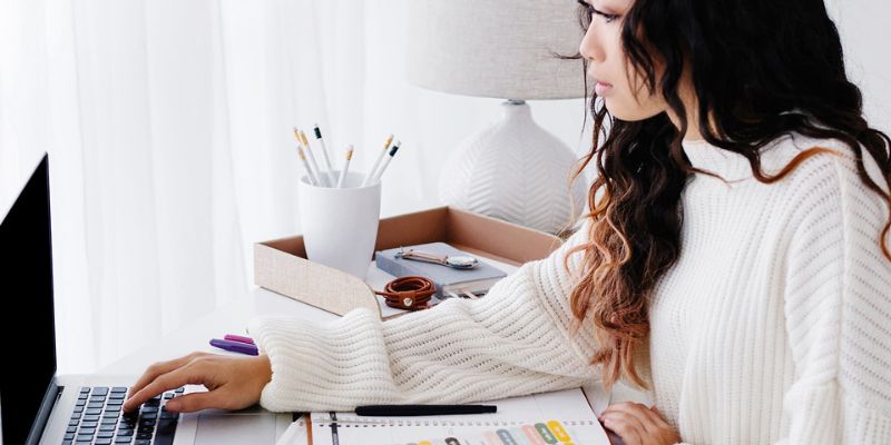 A person in a white sweater sits at a desk using a laptop, perhaps battling blogger burnout. Nearby, a notebook and colorful pens offer creative inspiration. A lamp and a cup holding pencils are in the background, completing the focused work or study environment.