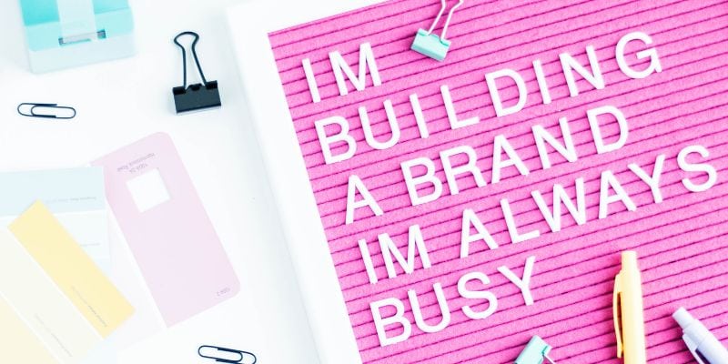 A pink letter board displays the message "IM BUILDING A BRAND IM ALWAYS BUSY" in white letters. Scattered office supplies, paper clips, and color swatches set the scene—perfect inspiration for chatGPT prompts for image generation.