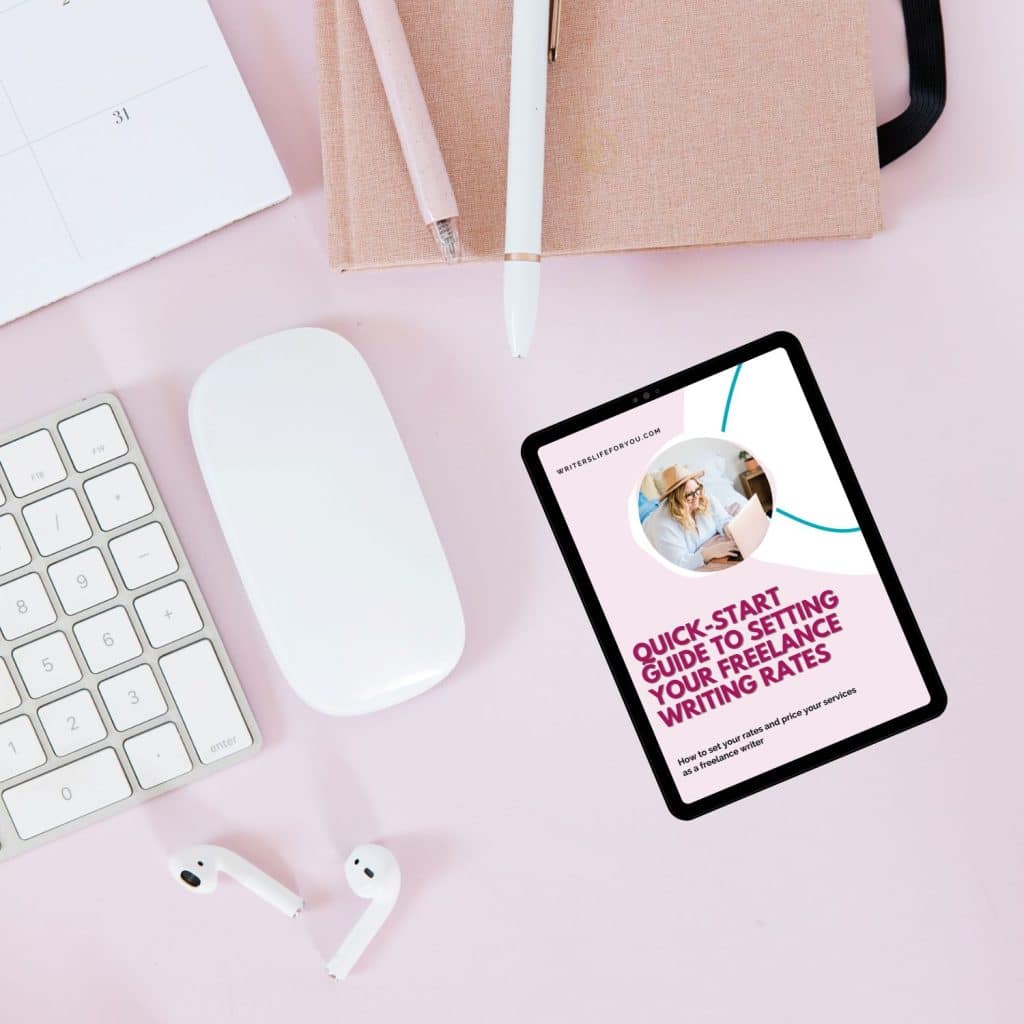 A desk setup featuring a white keyboard, wireless mouse, white earbuds, and a tablet displaying "Quick-Start Guide to Setting Your Freelance Writing Rates." The desk is pink, with a notebook and multiple pens placed on top.