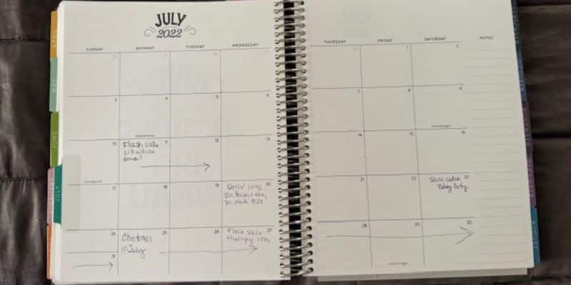 A spiral-bound planner is open to the monthly view of July 2022. The month is outlined with individual squares for each day, some containing handwritten notes. The notes include reminders about email, shipping orders, and a baby party. Tabs are visible on the left.