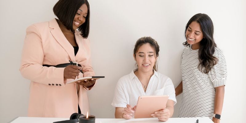 Three women are collaborating in an office setting. One woman in a pink blazer is taking notes on a clipboard, another in a white shirt is holding a tablet while seated, and a third woman in a striped shirt is standing and smiling. A lit candle on the table brings warmth as they discuss amateur blogging mistakes.