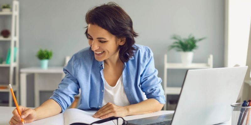 A person sits at a desk in a home office, writing in a notebook with a pencil. They are smiling, wearing a blue shirt, perhaps finding solace from blogger burnout as they sit next to an open laptop. The background includes shelves with a plant and other decorations.