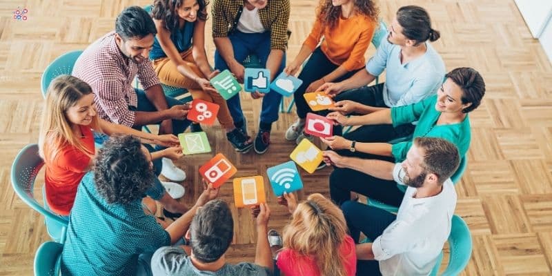 A group of people seated in a circle, each holding a square card depicting different social media icons such as Facebook, YouTube, Twitter, and Instagram. The wooden floor and bright light create a casual and collaborative atmosphere perfect for brainstorming social media content buckets.