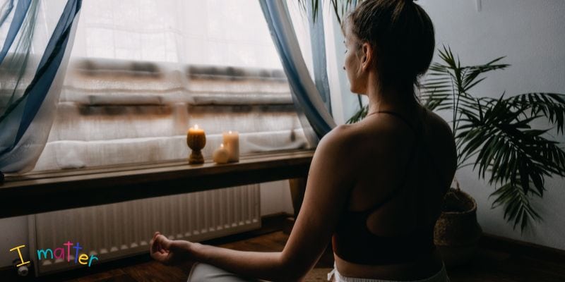 A person sits cross-legged on the floor facing a window with sheer curtains, seeking peace amidst blogger burnout. Two lit candles rest on a wooden bench beside the window, surrounded by potted plants. The words "I matter" appear in colorful letters in the bottom left corner, offering a gentle reminder.