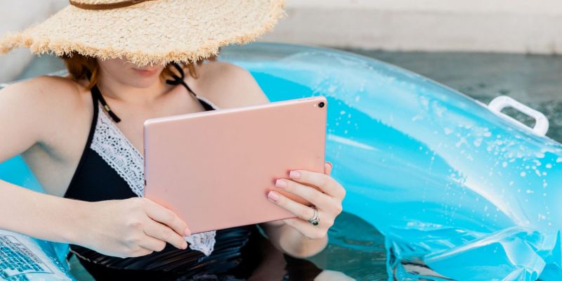 A person wearing a straw hat and black swimsuit is lounging in a blue inflatable pool float, holding a tablet. The scene is set for relaxed leisure and reading, offering the perfect escape from blogger burnout.