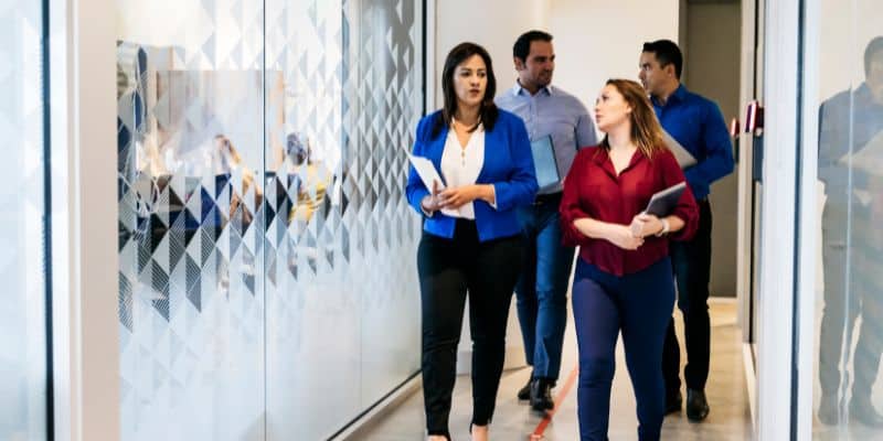 Four office colleagues, two women and two men, walking through a modern office hallway, carrying documents related to SEO blog writing services and engaged in conversation.