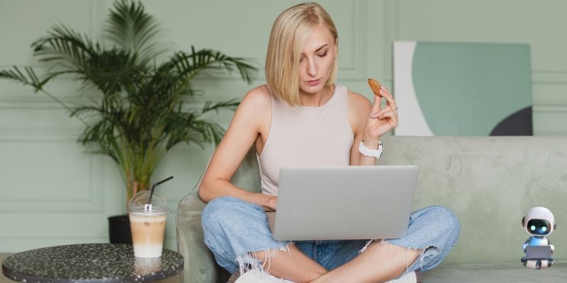 A woman sits cross-legged on a sofa, using her laptop to explore ChatGPT prompts for content writing while holding a cookie. An iced coffee is on a nearby table, with a small robot toy and a potted plant in the background.