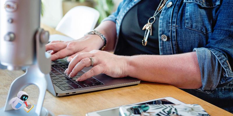 A person wearing a denim jacket types on a laptop at a wooden table, possibly working on ChatGPT prompts for content writing. A smartphone, microphone, and stationery are scattered nearby in this casual workspace.
