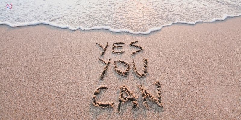 The image shows "YES YOU CAN" written in the sand on a beach near the shoreline, with small waves approaching the text—a perfect piece for your social media content buckets.