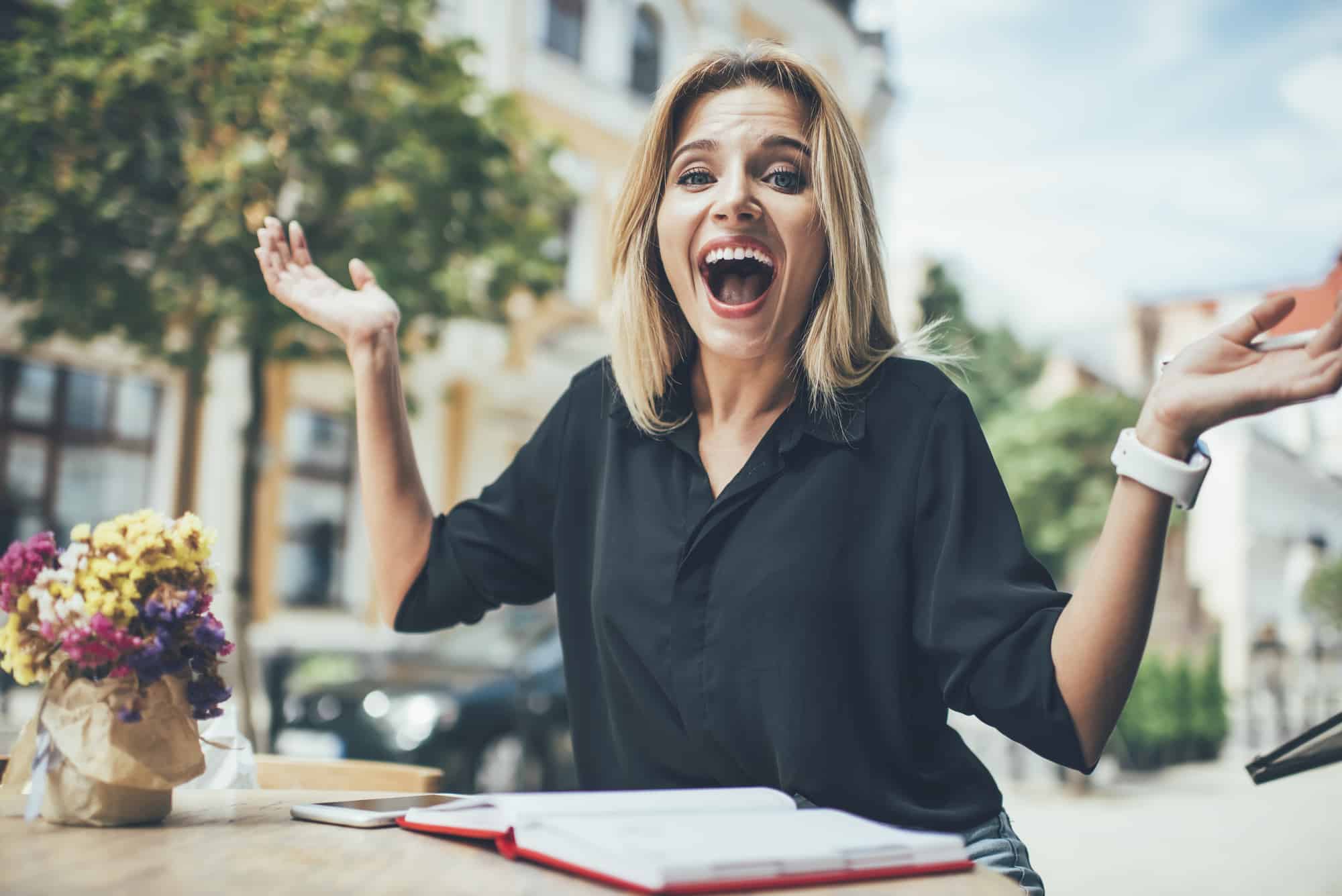 A woman with blonde hair, wearing a black shirt, sitting at a sidewalk table, expressing surprise or excitement with arms raised and mouth open wide. there are flowers and a closed book on the table.