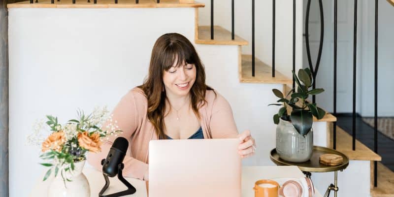 A woman sits at a desk with a microphone and laptop, smiling as she reads the screen. The room is bright with a staircase in the background and decorative plants around. She is preparing for her next