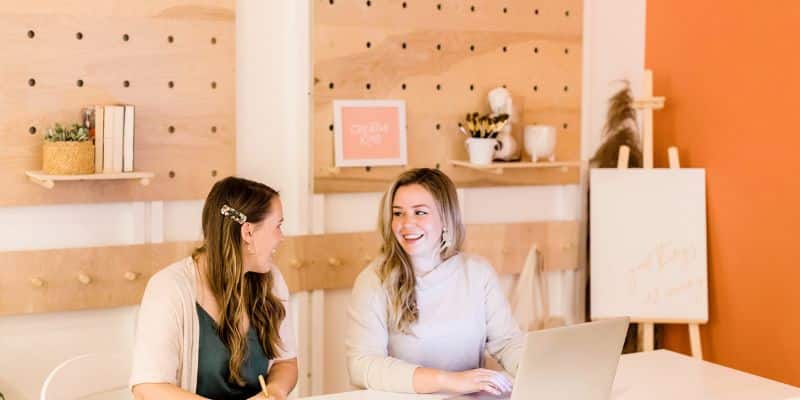 Two women smiling and engaging in conversation while sitting at a table with a laptop, discussing SEO blog writing services in a café-style setting with a modern, wooden wall rack and decorative items in the background.