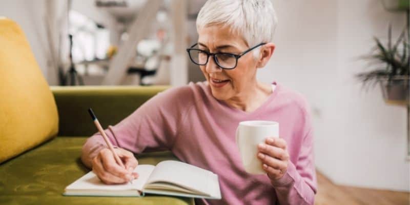 woman in pink shirt writing on couch with notebook and coffee