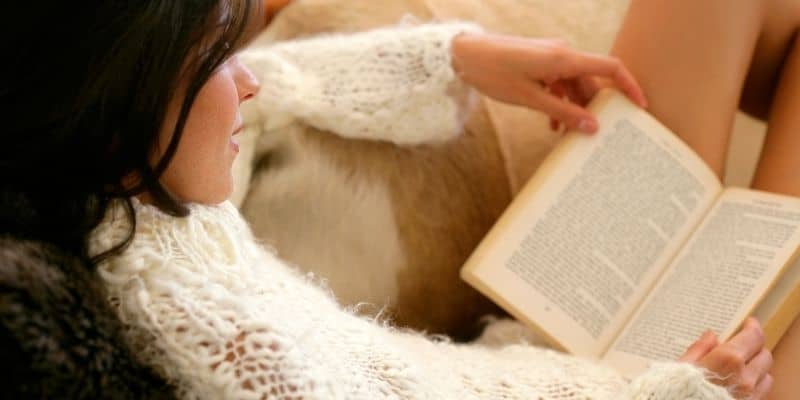 woman in sweater laying on couch reading book