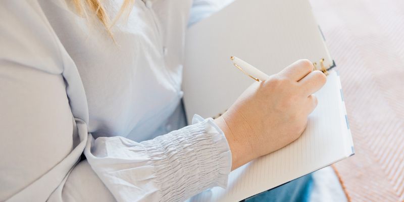 A person with light-colored hair is writing in a notebook using a pen, exploring the role of technology in writing. They are wearing a long-sleeved, light-colored blouse. The notebook is open on the table, and the background is blurred.