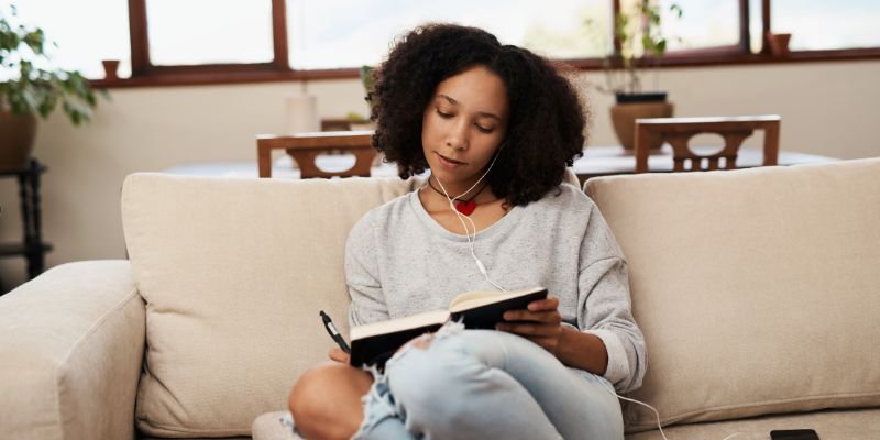 woman on a beige sofa listening to music while she journals