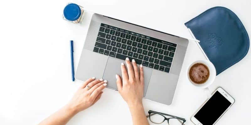 woman on laptop with white smartphone, blue pen, glasses, and cup of coffee