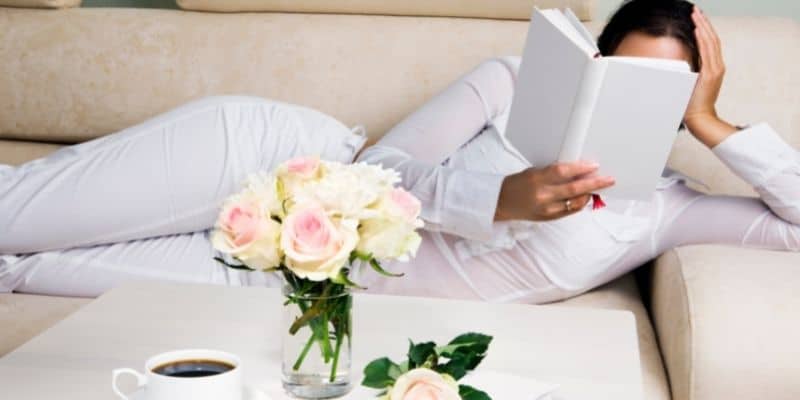 woman reading book on beige couch with flowers and coffee on table