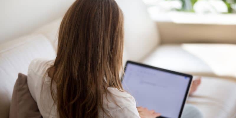 A woman with long brown hair is sitting on a couch, viewed from behind, looking at a digital tablet displaying an SEO blog writing services article.