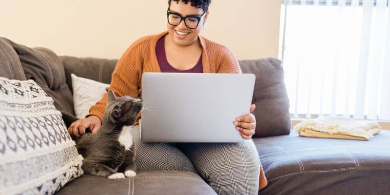 A person wearing glasses and an orange cardigan smiles while using a laptop for SEO blog writing services, sitting on a sofa next to a grey cat looking at the screen.