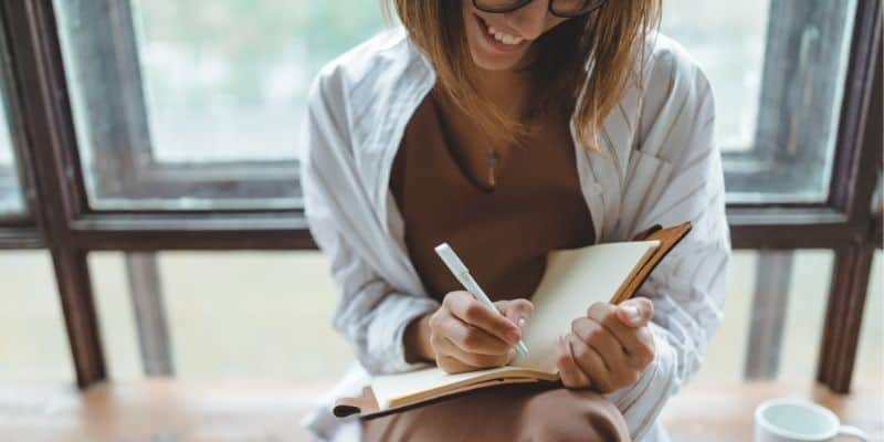 woman sitting in front of window writing with notebook and pen